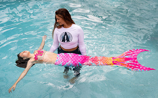 Woman helping girl in mermaid suit float in pool
